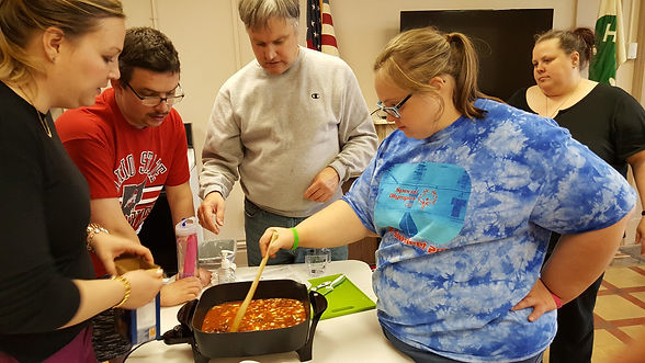 blonde adult woman stirring food in pan one adult woman, two adult men helping prepare food