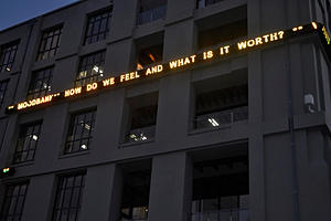 A lightboard on a building, it reads: how do we feel and what is it worth?