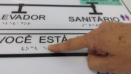 Exemplo de mapa tátil. Vê-se parte da palavra Elevador. À direita está a palavra Sanitário. Abaixo das palavras em tinta há a inscrição em braile. Um dedo está sobre um dos pontos pretos.