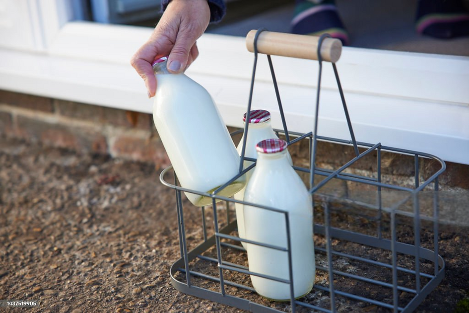 A hand retrieves a glass milk bottle from a metal crate on a doorstep