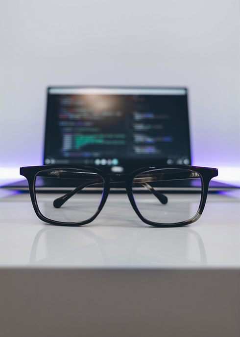 A pair of black-rimmed glasses sits on a white table, in front of a blurred laptop screen displaying colorful lines of code