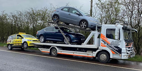 Tow truck carrying two cars, one silver and one blue, on a rainy roadside
