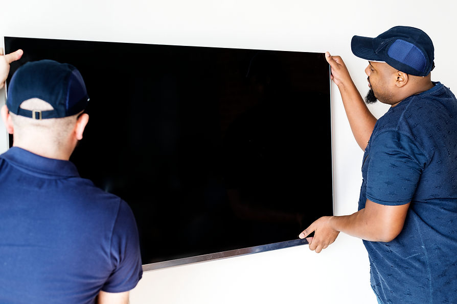 Two men in blue shirts and caps are mounting a large flat-screen TV on a white wall