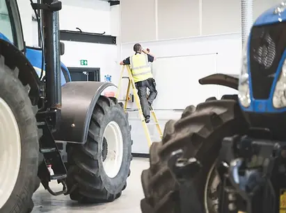 Two tractors indoors with a worker climbing a ladder in the background