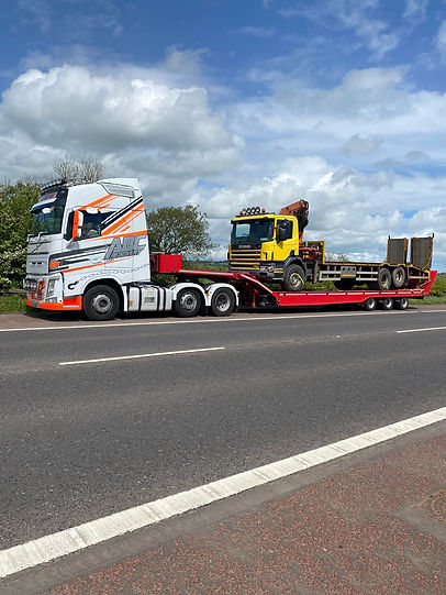 A white and orange truck tows a flatbed trailer with a yellow truck cab on it