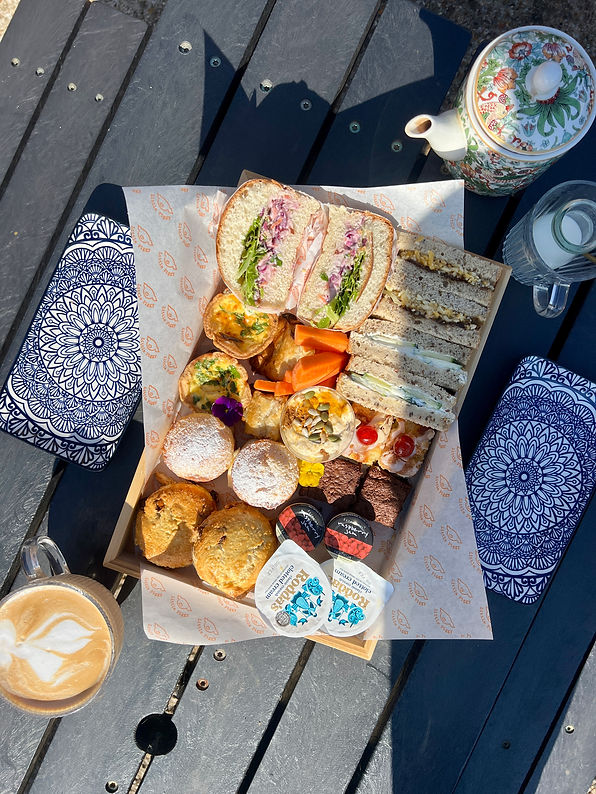A wooden tray on a table holds assorted sandwiches, scones, pastries, and condiments