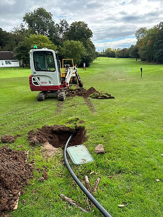 An excavator digs a trench on a grassy field, revealing a buried cable