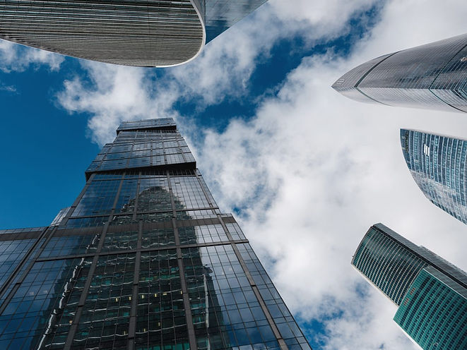 Skyscrapers viewed from below against a blue sky with clouds, reflecting each other. Modern architecture, glass facades, and an airy atmosphere.