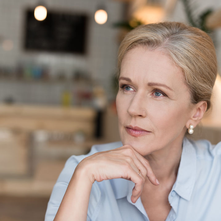 A middle-aged woman sits in a café, chin resting on her hand, looking thoughtfully into the distance — reflecting on how much of her portfolio should be in stocks