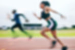 Two athletes sprint on a track, one in focus wearing green and black, the other blurred.