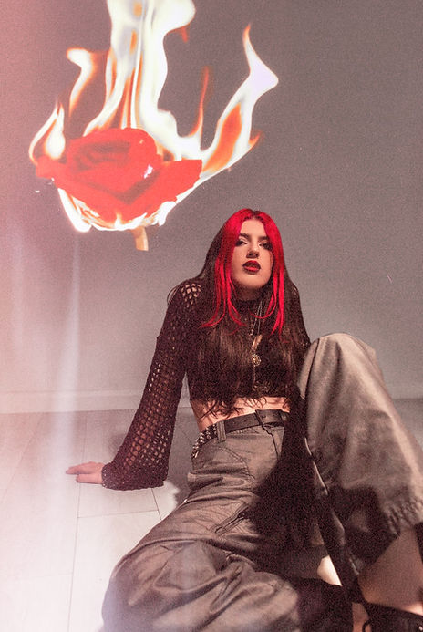 Edgy photo of a red haired girl with a rose on fire behind her, dark makeup and red lips.