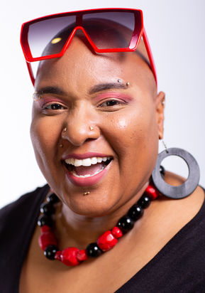 Sharon smiles at the camera with oversized red sunglasses lifted above her eyes, a large black hoop earring, and a red and black beaded necklace. She has a shaved head and looks to be mid-laugh.