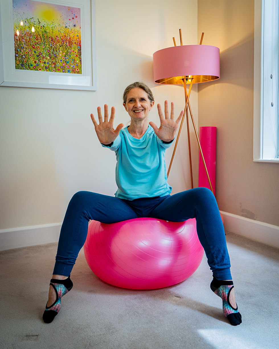Kerry wearing a turquoise top and blue trousers, sitting on a pink stability ball smiling and with her hands stretched out in front
