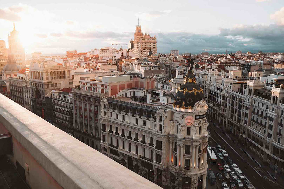 Cityscape of Madrid at sunset, featuring the Metropolis Building with a dark domed roof. The sky is partly cloudy, casting a warm glow.