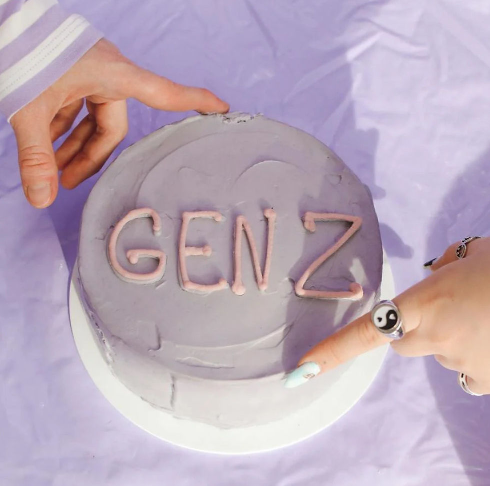 Hands touch a purple cake with "GEN Z" in pink icing. One hand has a yin-yang ring. Background is a matching purple cloth.