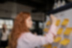 Woman with red hair writes on flip chart labeled "Marketing" covered in yellow sticky notes. Office setting, focused expression.