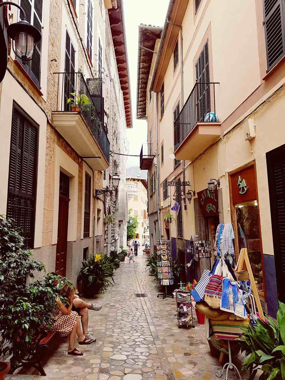 Narrow cobblestone alley with a person sitting on a bench, surrounded by plants. Boutique store displays bags and scarves. Cozy, vibrant mood.
