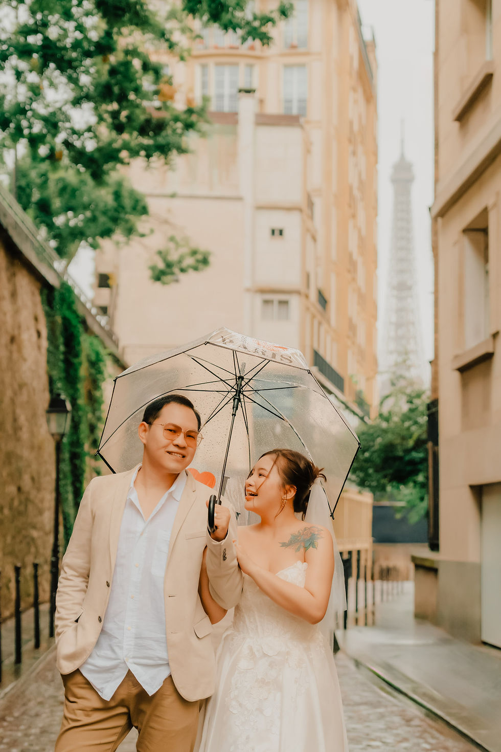 A couple strolls through the charming streets of Passy, sharing an umbrella on a rainy day, with the Eiffel Tower subtly peeking in the background. Photo by lovidovi.art