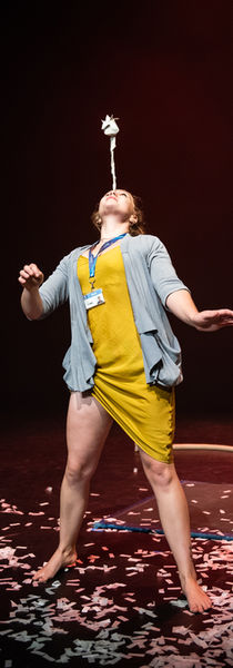 performer in yellow dress balancing a paper rose on her nose while standing on a stage covered in white confetti