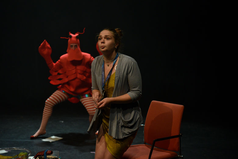 circus performer in yellow dress with lanyard around her neck is getting out of a chair while behind them is a performer in red lobster costume