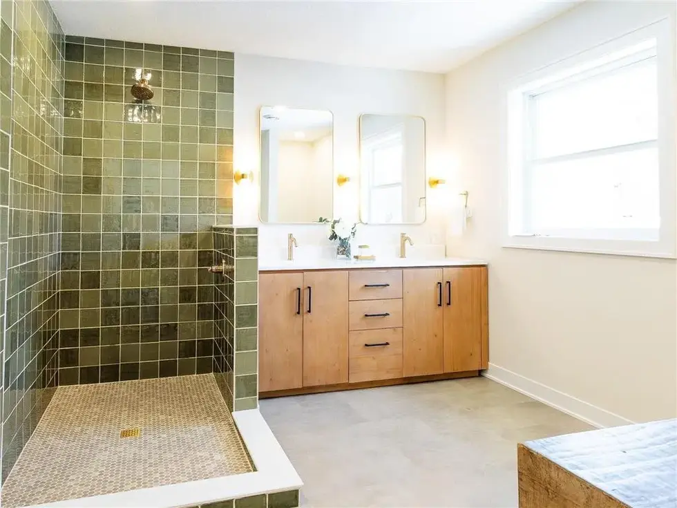 Dual sink vanity with gold fixtures and sconces placed on each side of the mirrors with a green square tiled shower
