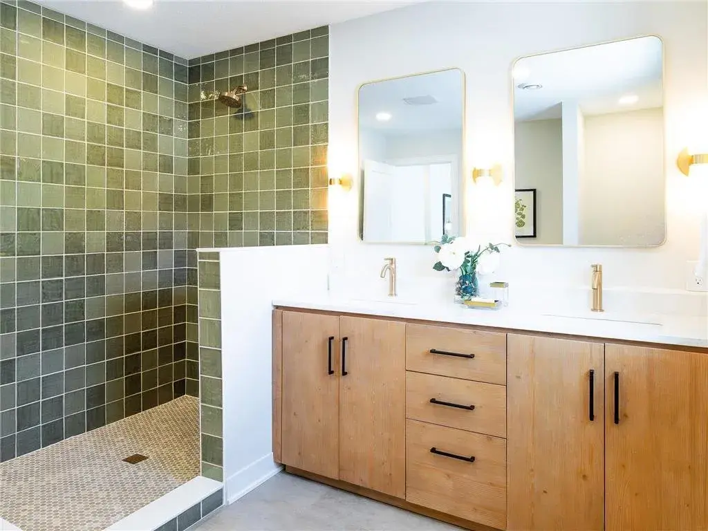 Dual sink vanity with gold fixtures and sconces placed on each side of the mirrors with a green square tiled shower