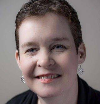 White woman with very short dark hair, wearing a black cardigan with a bold blue shirt underneath