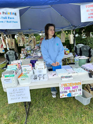 A girl stands behind a stall selling cards and advertising face painting and a tombola. 
