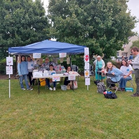 Children and adults man a stall under a blue gazebo on Furzedown. The stall is decorated with drawings, and sells cards and tote bags.