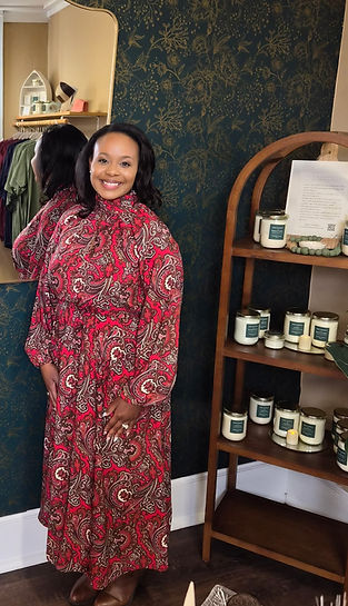 A woman in a red, ivory, and brown paisley dress is standing next to a shelf in a store, smiling.