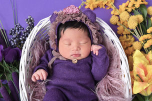 Beautiful baby girl in dark purple pajamas and flower bonnet laying in white basket surrounded by dark purple and yellow flowers. Bold and bright newborn photography at Key West photography studio. 