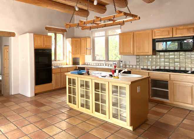 Open concept Santa Fe kitchen prior to remodel with Saltillo tile flooring, aged wooden cabinetry, rustic pot rack, and tile backsplash