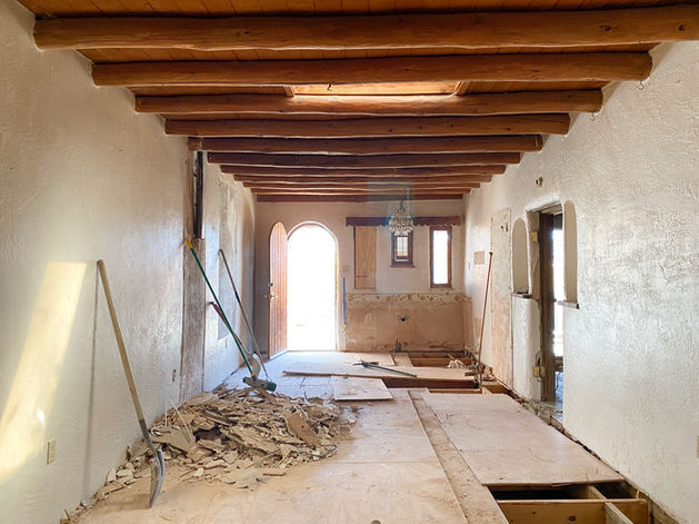 View through a bedroom space being remodeled with plaster walls, holes in the floor, swept debris, and an open arched doorway at the end of the room.