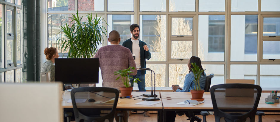 Four people in a bright office, engaged in a discussion. One stands speaking, others seated. Large windows, plant, and desks visible.