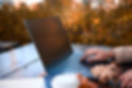 Hand typing on a laptop outdoors, surrounded by autumn leaves and a coffee cup, on a wooden table with a blurred foliage background.