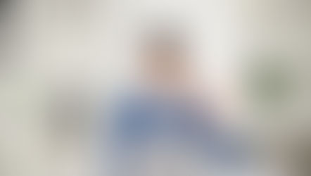 Man in blue suit smiling and waving at laptop in bright office. Bookshelf with decor in background. Casual, friendly video call atmosphere.