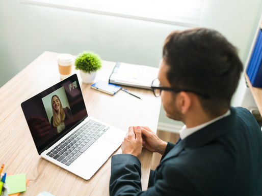 Man in a suit video calls a smiling woman on a laptop in a bright office. Desk has a plant, coffee cup, and notebooks. Mood is professional.