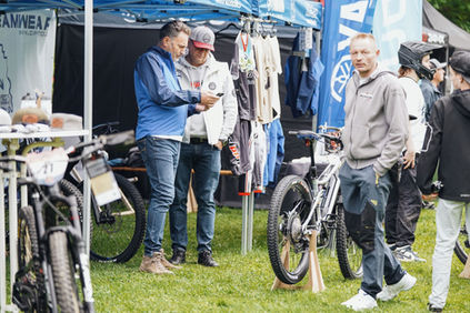 Drei Männer im Gespräch, vor einem Zelt, mit Fahrrädern und Textilien.