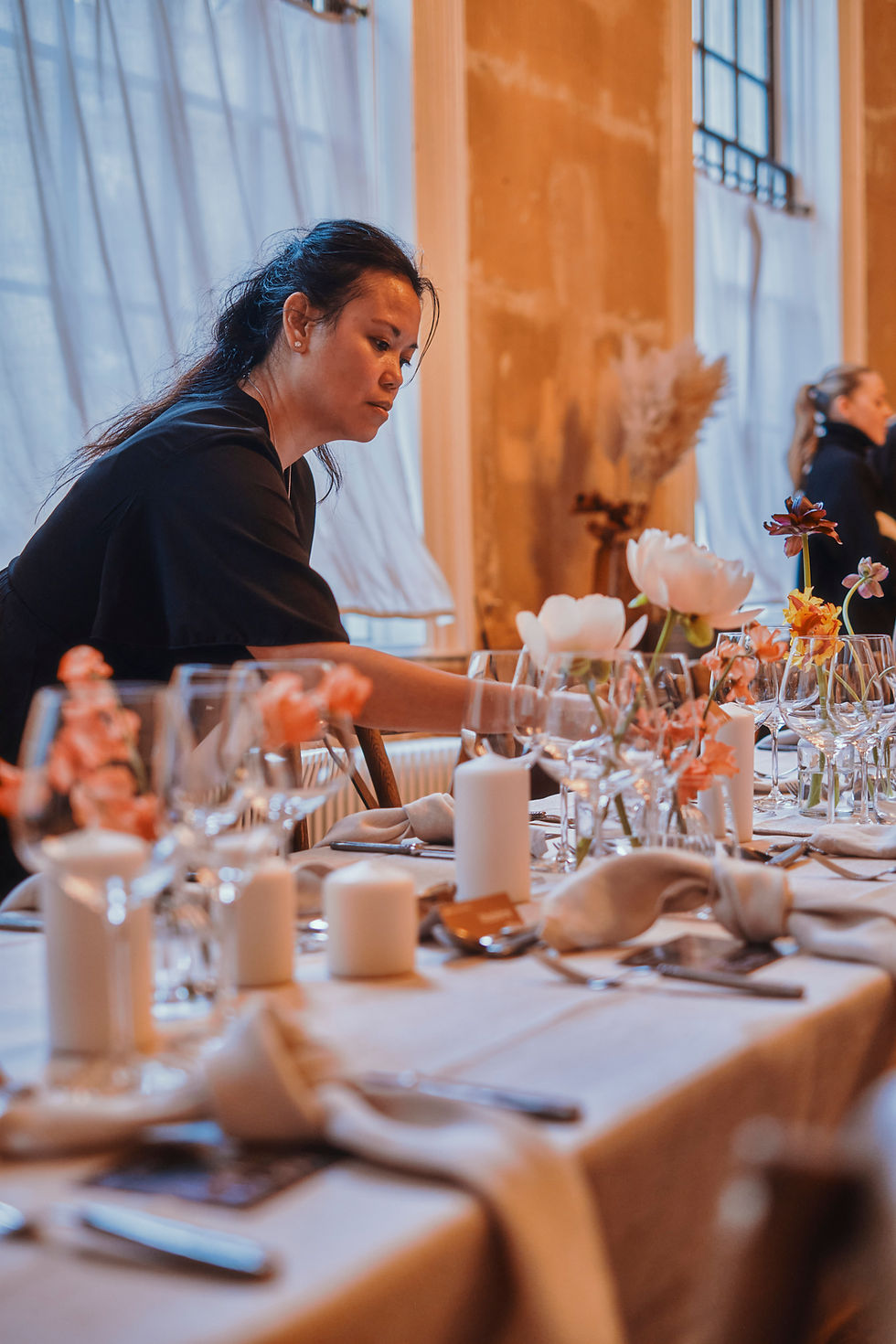 A woman sets a table with candles and flowers in a warmly lit room. Another person is visible in the background, creating a cozy atmosphere.