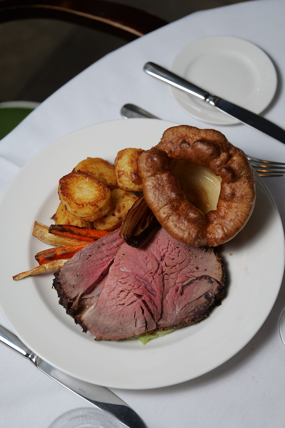 Roast beef with Yorkshire pudding, roast potatoes, and carrots on a white plate. Table setting includes a fork, knife, and small plate.