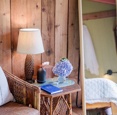 Heart Room at Hamakua Sanctuary featuring a cozy bedside setting with a wicker chair, lamp, essential oil diffuser, blue journal, and hydrangeas—designed for grounding and warmth