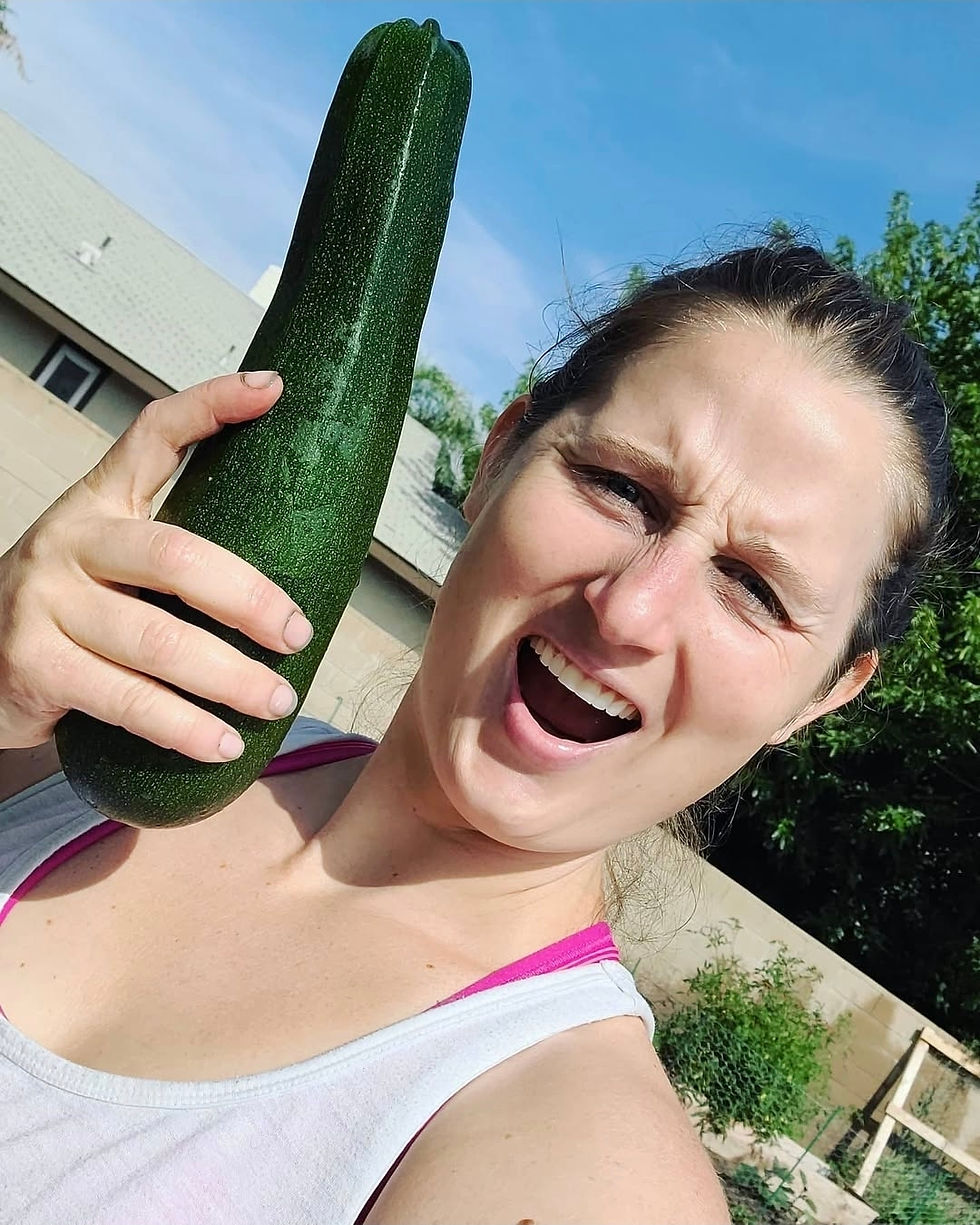 Woman in a sunny backyard holds a giant zucchini beside her face, smiling playfully.