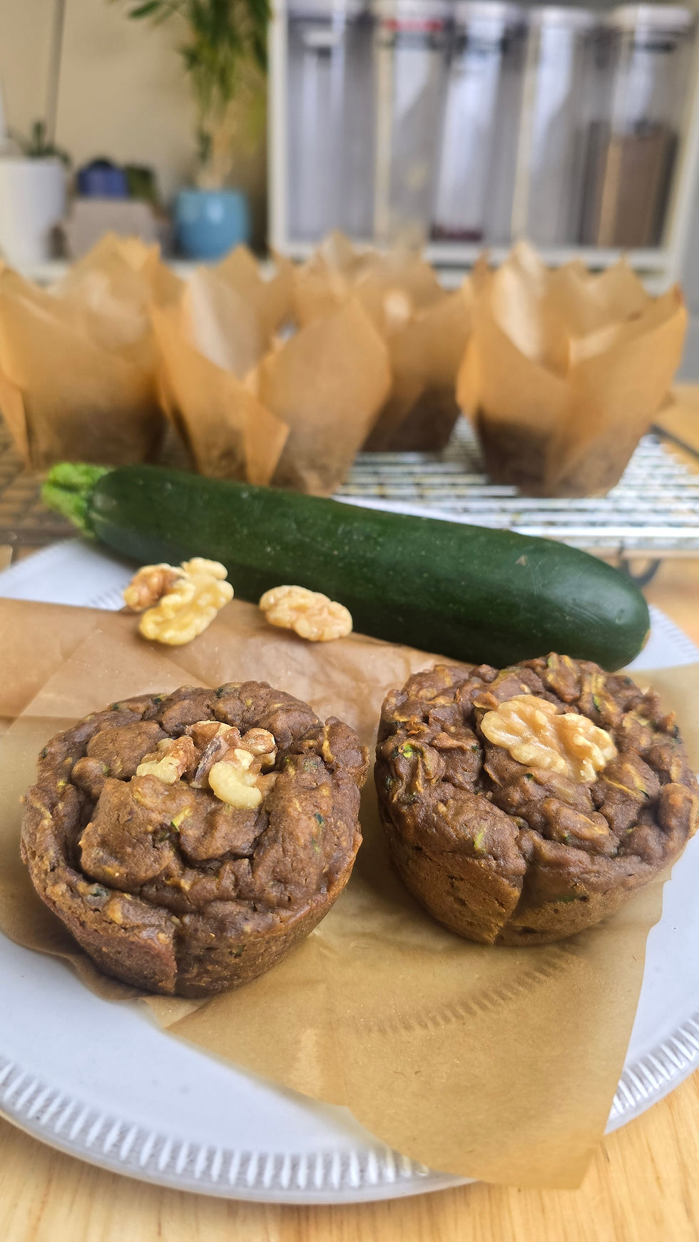 Two walnut-topped zucchini muffins on a paper plate with a whole zucchini behind them, baking setup in a cozy kitchen.