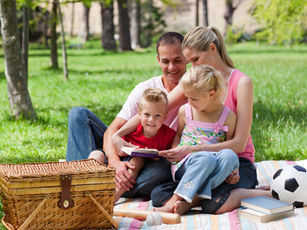 Family of four having a picnic on a blanket in a sunny park, reading together. A soccer ball, books, and picnic basket are nearby.