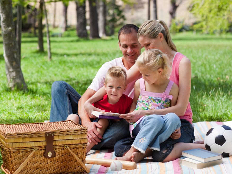 Family of four having a picnic on a blanket in a sunny park, reading together. A soccer ball, books, and picnic basket are nearby.