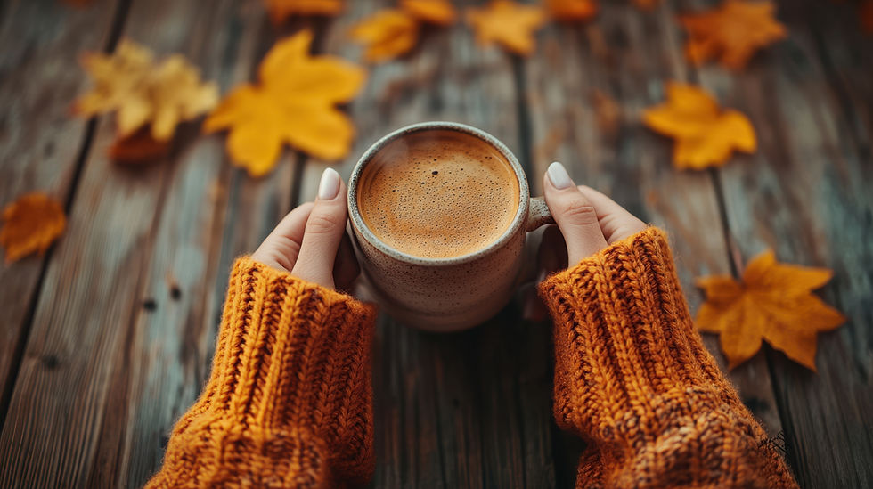 Hands in an orange sweater hold a steaming coffee cup over a rustic wooden table with scattered autumn leaves.