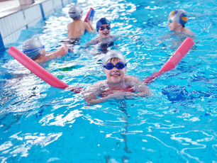 Children swim in a pool with colorful noodles, wearing caps and goggles. One child smiles in the foreground. Bright, lively atmosphere.