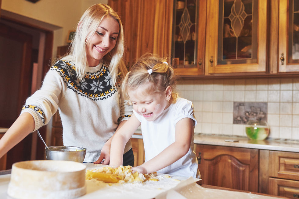 Smiling woman helps a young girl knead dough in a cozy wooden kitchen, with flour, bowls, and warm light around them.