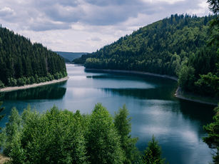 A serene river winds through lush green hills under an overcast sky, reflecting the surrounding forest. The mood is peaceful and calm.