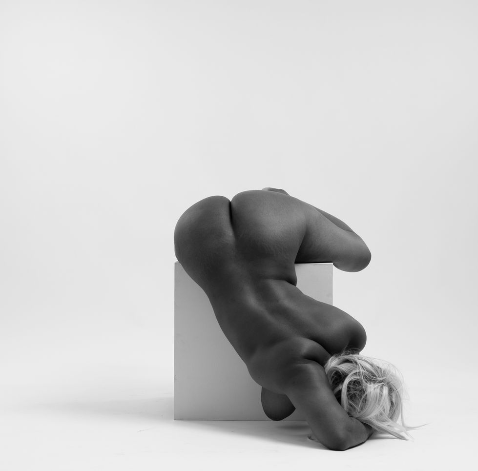 a black woman drapes her naked body over a white block against a white backdrop in this black and white photo."
